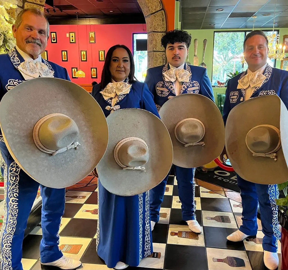 Cuatro integrantes del mariachi con traje azul bordado y sus instrumentos frente a una pared naranja