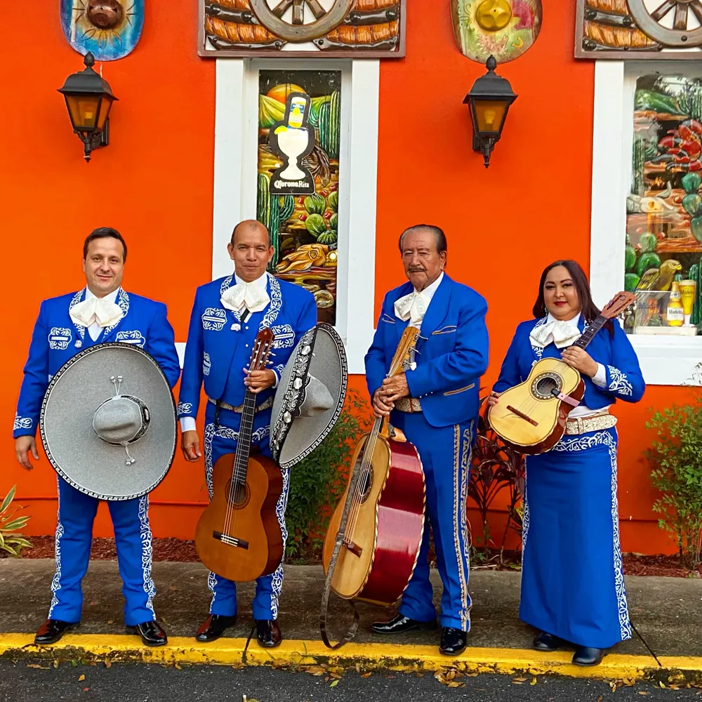 Cuatro integrantes del mariachi con traje azul y bordados claros sostienen sombreros grises en un restaurante