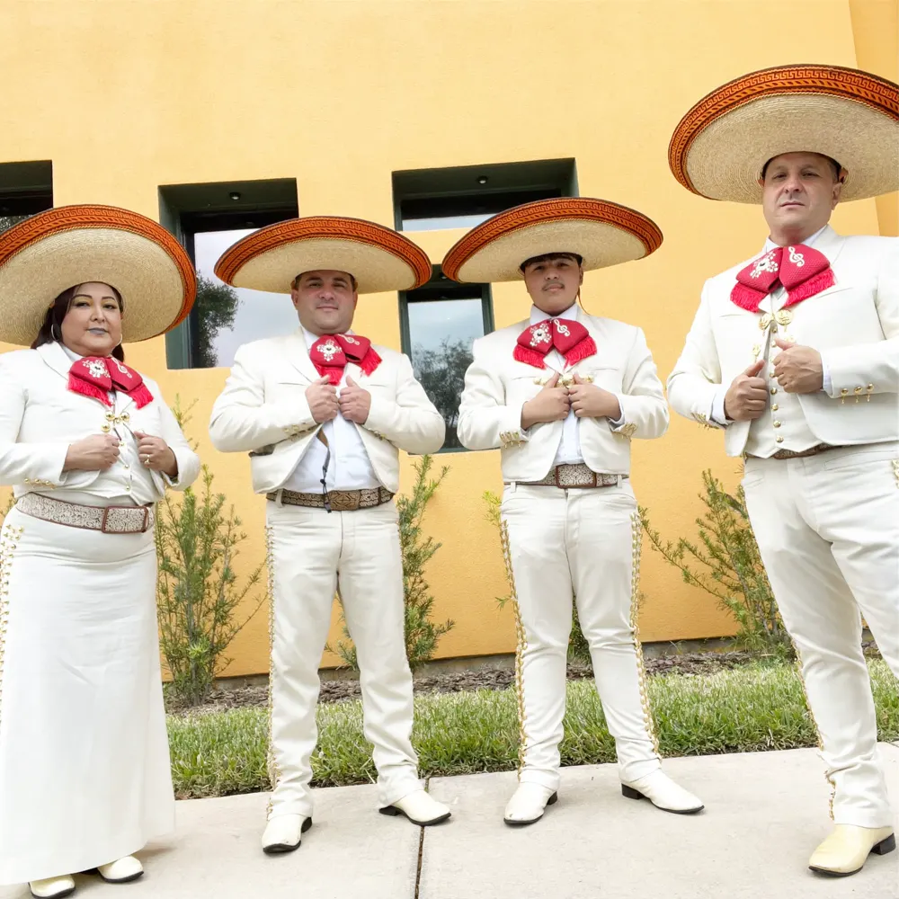 Cuatro integrantes del mariachi con traje blanco y moño rojo, con sombreros de ala ancha, frente a una pared amarilla