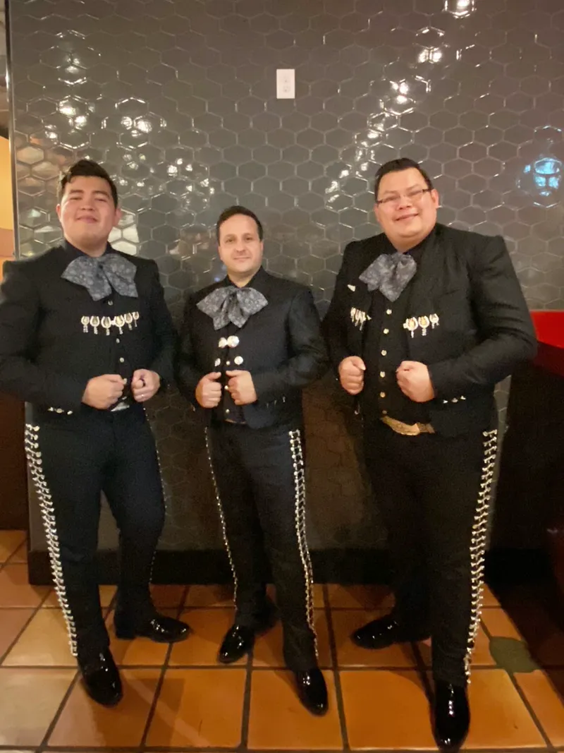 Tres músicos del mariachi de pie, con traje de charro negro adornado y moño gris, posando para la foto