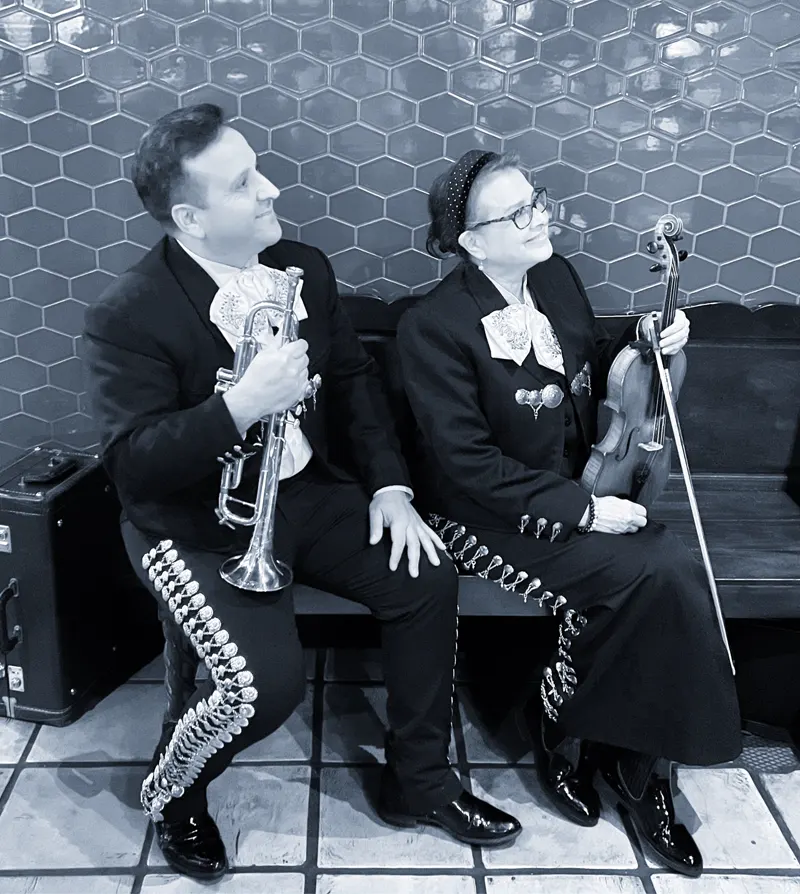 Foto en blanco y negro de un trompetista y una violinista del mariachi sentados frente a una pared de azulejos